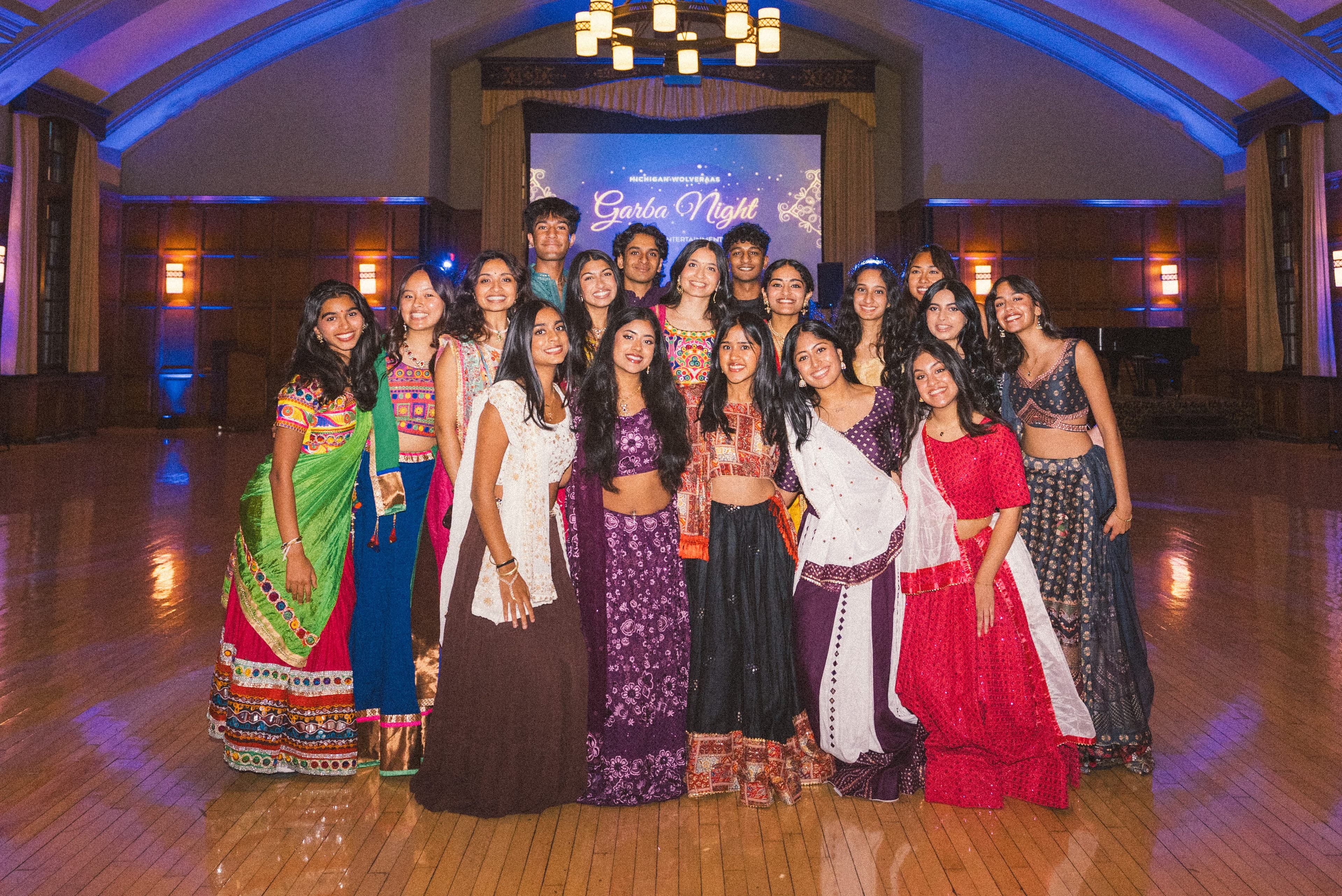 A large group in colorful traditional outfits posing in front of the Garba Night screen