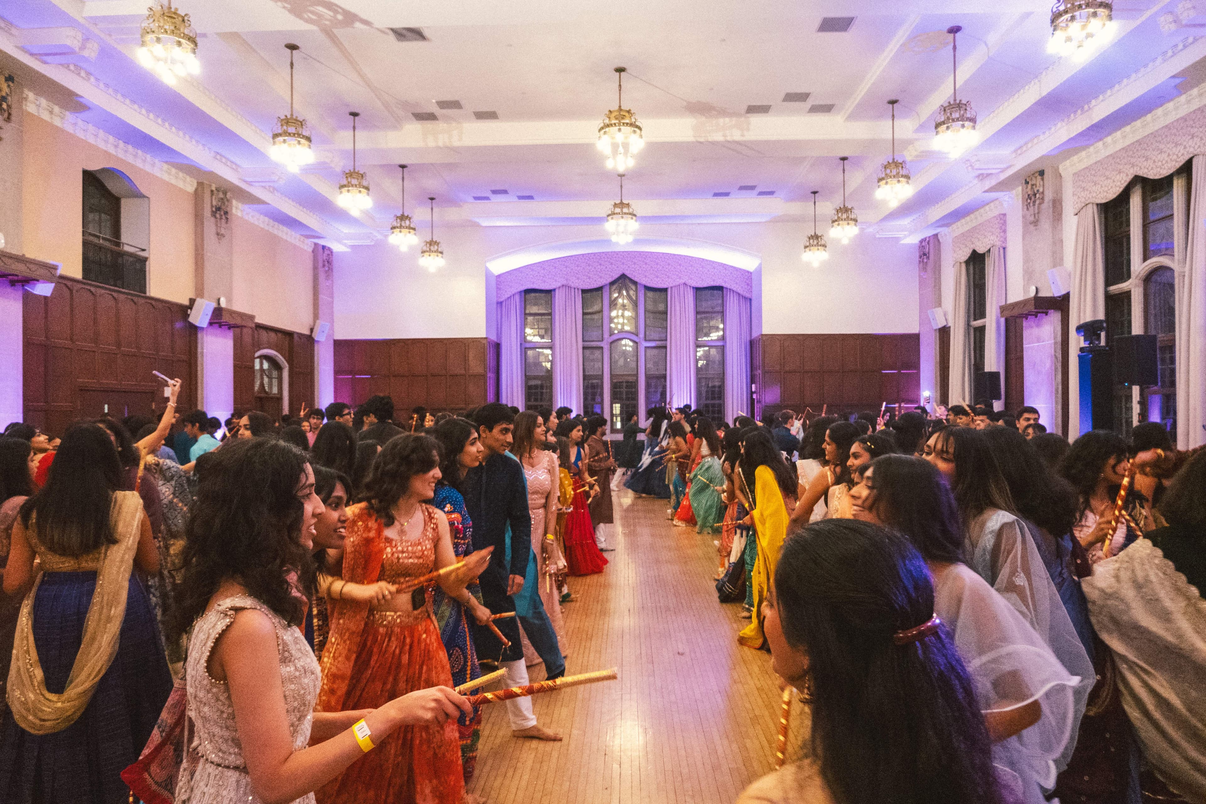 A packed dance floor of Garba dancers under purple light, seen from a balcony above