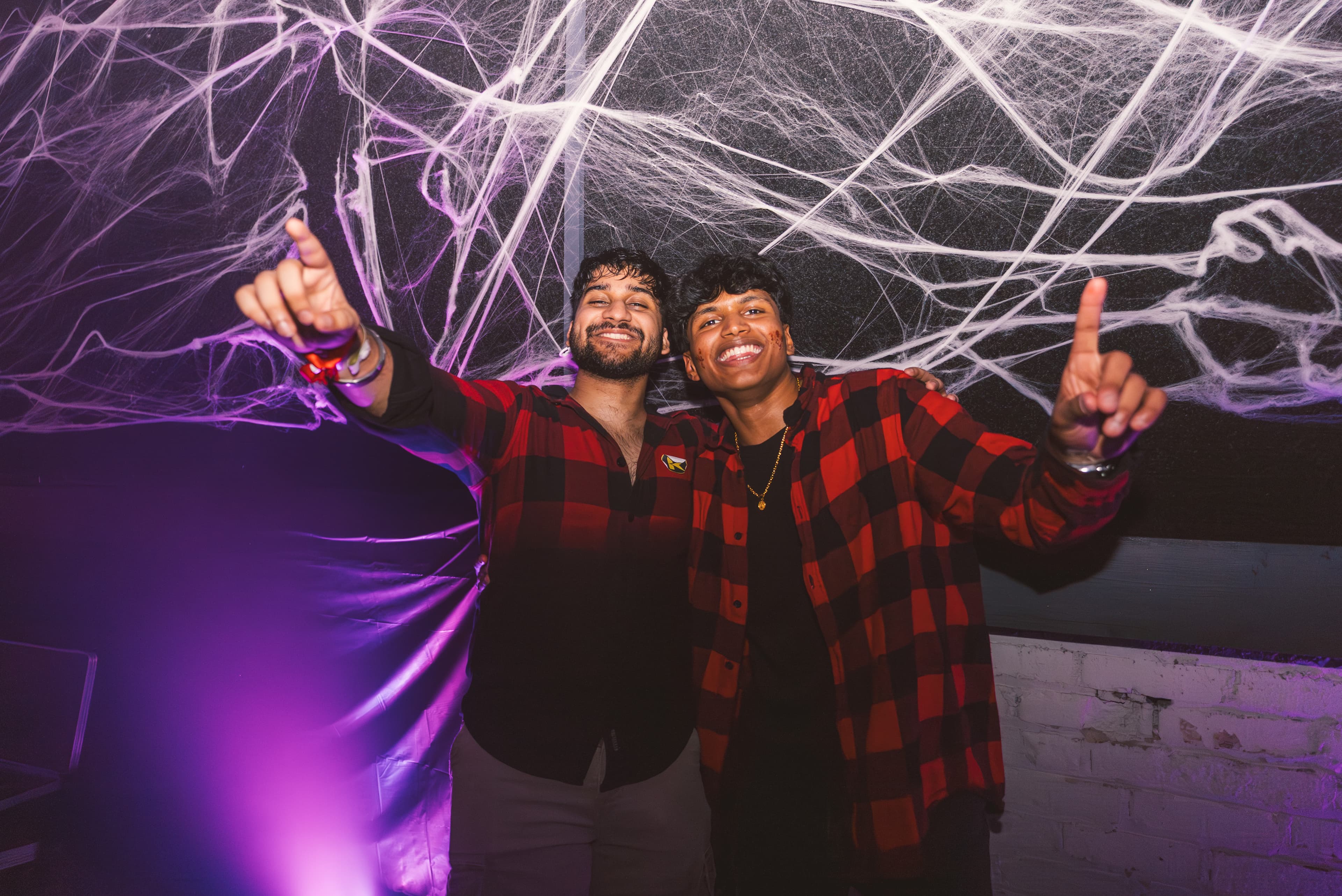Two friends in matching red flannel shirts pointing up under purple cobwebs